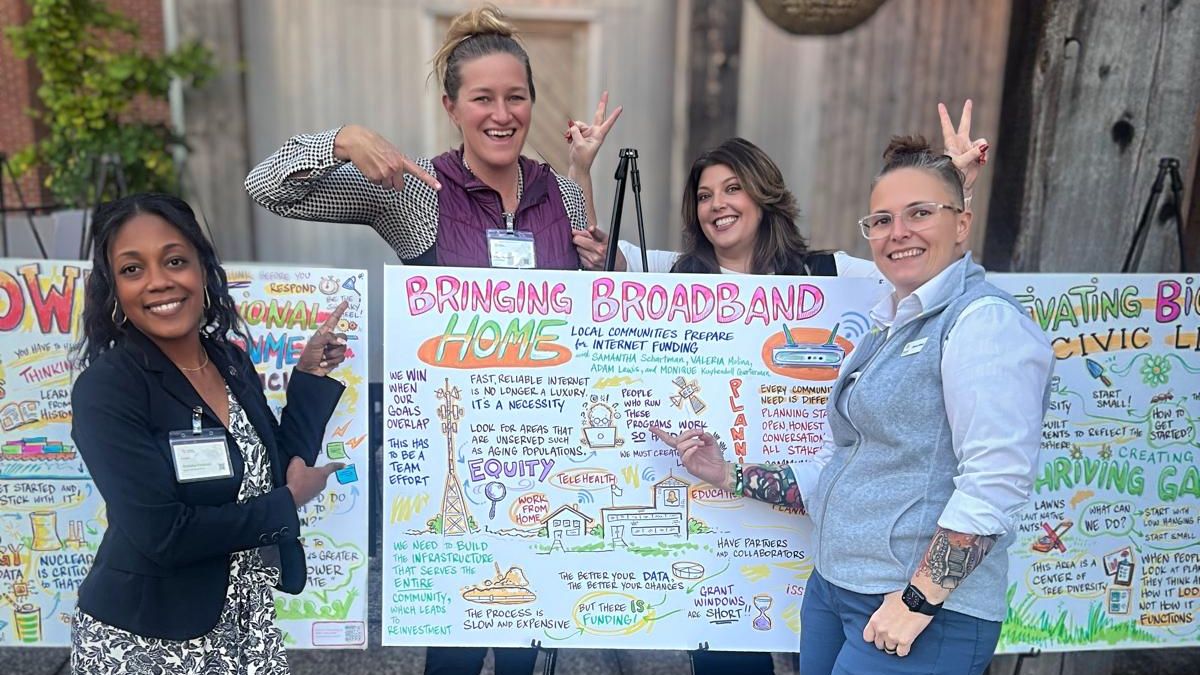 Photo of four women standing at board that says "bringing broadband home"
