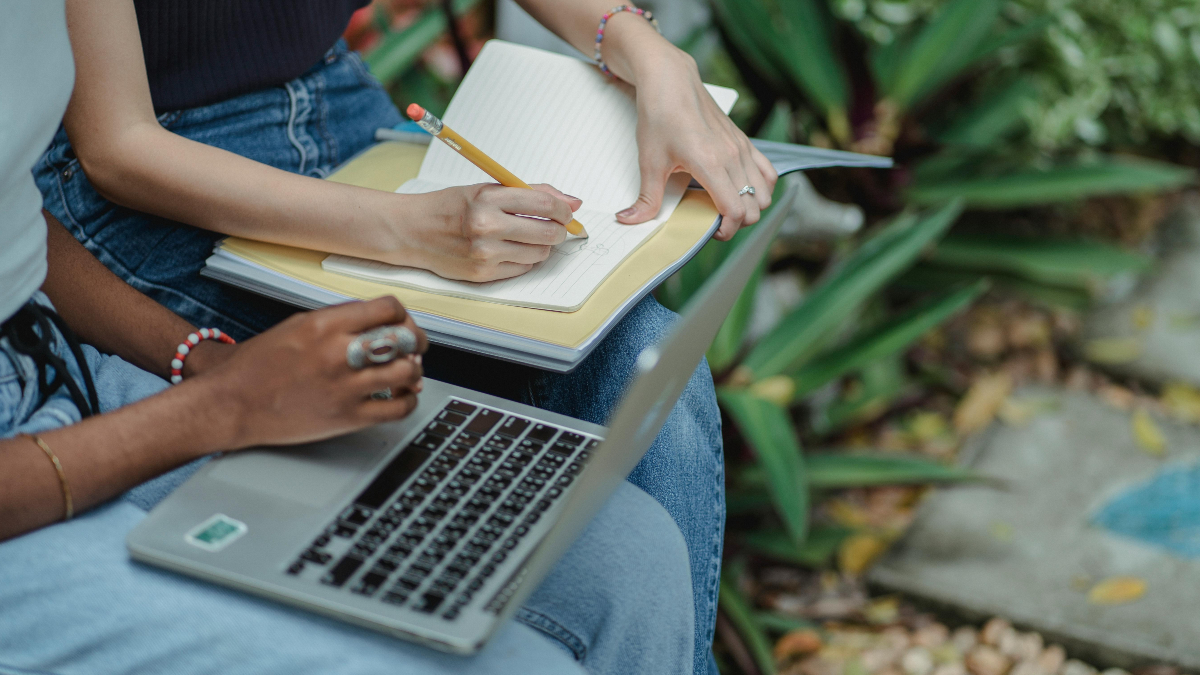 Two school aged students on bench with laptop and notepad